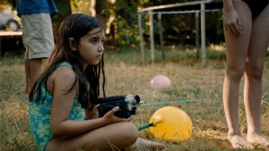 Eylul Guven as Sasha sitting on the grass in a yard holding a camera in a still from "Blue Heron."