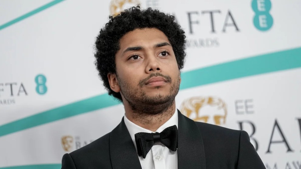 A man with dark-toned skin stands in front of a step-and-repeat background, wearing a tuxedo, with a solemn look on his face.